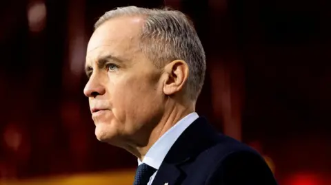 A side profile image of Mark Carney from the shoulders up. H ehas short black and grey hair, and is wearing a black suit and patterned tie. Behind him is a blurred out dark burgundy background. 