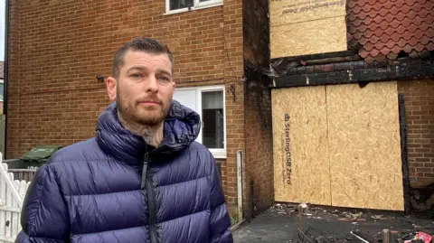 Dean Ray looking into the camera with a slight smile. He has brown, short hair which has been gelled to the side on top. He is wearing a blue puffer coat and has tattoos on his neck which are just visible. He is standing in front of the burnt house which has cardboard covering the door and window above it. There is rubble on the floor.