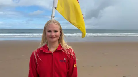 Freya Macfarlane has long blonde hair and is wearing a red RNLI lifeguard fleece. She is standing on Whiterocks beach in front of a red and yellow lifeguard flag.
