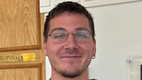 James Turner/LDRS Niall Walker has short black hair and a beard. He is wearing rim-less glasses and a hoop earring in the right ear. He is standing in a corridor, behind him a a wooden on the left with a gold plaque reading 'CHAMBER'.