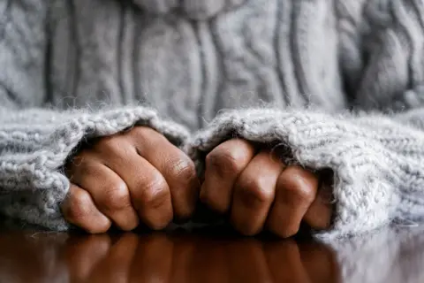 A stock image of hands in fists, resting on a wooden surface. The person is wearing a grey woolly jumper and its sleeves are pulled up close to the person's knuckles. 