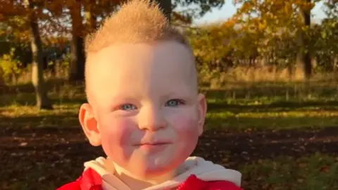 Family/GoFundMe A boy with brown hair and a red and white top is looking at the camera. Grass and trees are behind him.