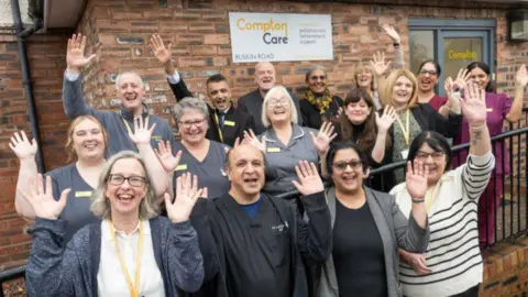 A group of 16 people stand and wave at a camera, outside a redbrick building with a white sign on the wall saying "Compton Care", with Compton in yellow. Many of the people have similar-looking grey scrubs with the same yellow on pin badges attached to their uniforms, with others wearing yellow lanyards.