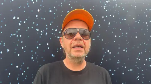  Llyr Ifans is wearing sunglasses, an orange cap and a black T-shirt