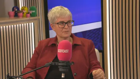 BBC Naomi Cunningham sitting in a BBC podcast studio. She is behind a microphone with a red cover that says Scotcast. She has short, grey hair and glasses. She is wearing a red jacket