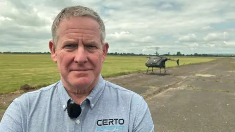 The CEO of a helicopter manufacturer stands in front of his unmanned helicopter at Keevil Airfield.

