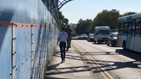 Shariqua Ahmed/BBC A woman in white shirt and trousers, is walking on a footpath with two people on an e-bike coming her direction.