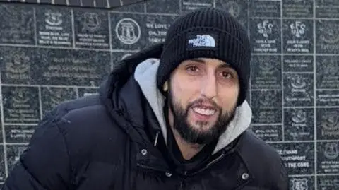 A man with a dark beard smiles at the camera. He wears a black North Face hat and a black coat with a grey hoodie underneath. 