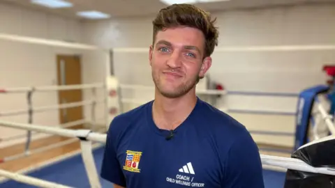 Stuart Woodward/BBC Joel Brown wears a navy blue T-shirt with a club badge on one side of his chest for the Southend Amateur Boxing Club, and on his other side is an Adidas logo and his title of coach/child welfare officer. Joel has short brown hair and a stubbly beard. He is standing in front of a boxing ring.