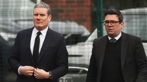 Starmer and Burnham walk alongside one another both in suits in front of a gate with cars behind it as they attend the funeral of Sir Tony Lloyd in 2024.