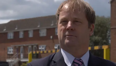 A man with light brown hair who is wearing a blue suit, light blue shirt and pink and blue tie. He is standing in a car park with red-brick buildings and a yellow and black entrance barrier in the background.