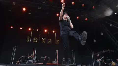 Sharleen Spiteri from the rock band Texas. She is wearing a black top and jeans and black boots. She has black hair. She is stood on stage holding a microphone to her mouth with one hand up in the air. Her band mates can be seen behind playing guitars and drums.