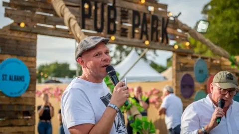 Pub in the Park Chef Tom Kerridge wearing a light grey T-shirt and darker grey flat cap, holding a microphone in front of a sign saying Pub in the Park in St Albans