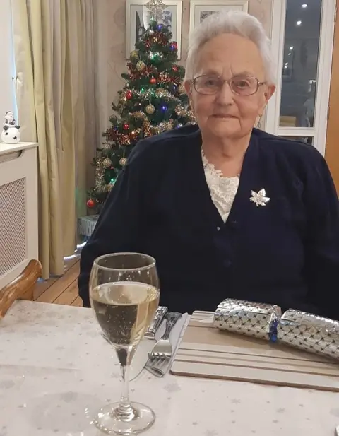 An elderly woman with short grey hair and glasses sits at a table with a glass of wine and a Christmas cracker 