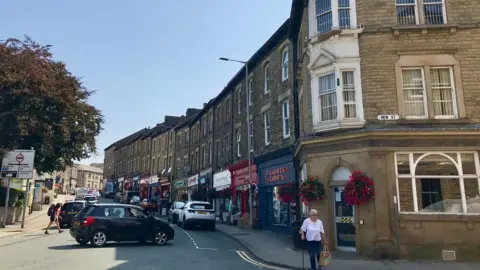 LDRS Carnforth town centre with cars in the middle of the road and a row of shops in distinctive quarried stone on the right hand side.