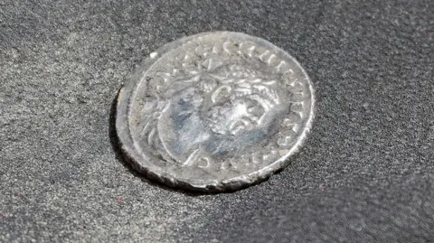 A close-up photograph shows an aged silver coin resting on a dark textured surface. The coin features a worn profile portrait surrounded by an inscription around the edge, although much of the detail has been softened over time. Visible wear and slight damage along the rim suggest the coin has been buried or handled extensively. The image highlights the coin's historical character and archaeological significance.