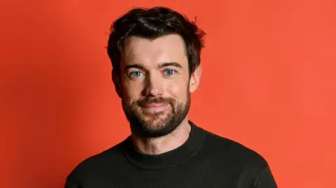 BBC Jack Whitehall, wearing a black shirt with a round collar, stands in front of a red backgroumd. He has dark brown hair, a beard and blue eyes.
