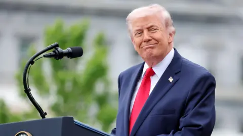 PA Trump standing at an outdoor podium with a microphone, wearing a dark suit and red tie, with blurred greenery in the background.