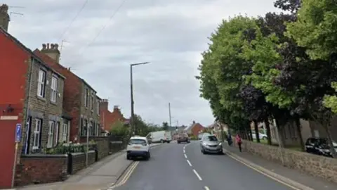 A road with terraced houses on one side and a row of tress on the other side. Pedestrians walking on the pavement.