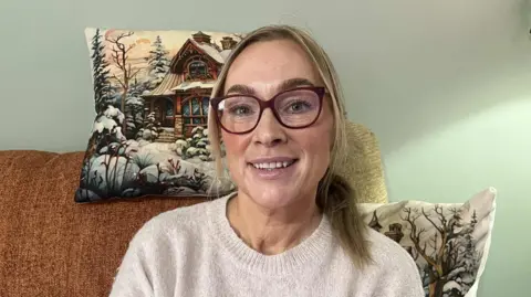 A woman wearing a stone-coloured jumper. She is sitting on a couch with a light brown blanket. Behind her head is a Christmas themed cushion and her left-hand shoulder is another Christmas themed cushion 