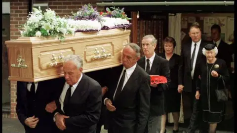 PA Media Mourners dressed in black carry a light wood coffin adorned with floral tributes on top
