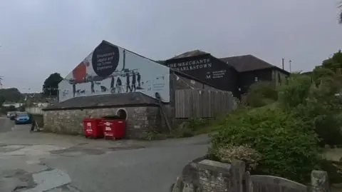 Google Google Street View image of the Shipwreck Treasure Museum in Charlestown. On one gable wall is a mural depicting an exhibition on Ernest Shackleton. Directly below the mural are two large red containers.