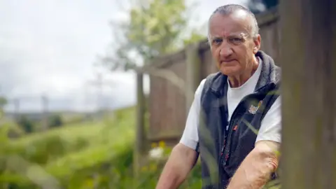 Rogan Scotland William Grant is looking at the camera with a serious face. He has cropped, grey hair and is wearing a white T-shirt with a blue fleece gilet over the top.  He is leaning forward on his arms and there is a wooden building and fields behind him.