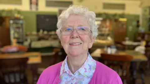 Margaret Lees is smiling at the camera. She has short grey hair and is wearing silver glasses. She is wearing a pink cardigan and a white blouse.