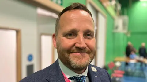 Councillor Matt Dormer is pictured - he is inside a leisure centre building wearing a blue suit jacket and a green shirt, with a lanyard around his neck. 