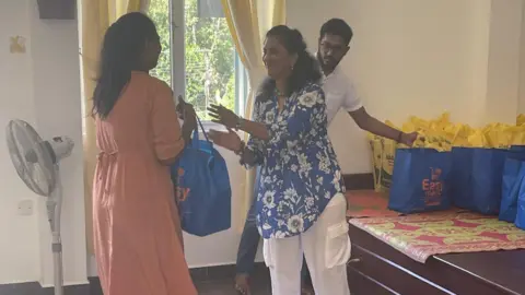 Supplied A woman in a blue floral top passing a blue bag to someone else with a smile on her face. Behind her is several identical bags.