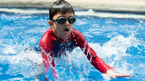 Getty Images A young boy wearing a red wet suit and black goggles splashes in a swimming pool.