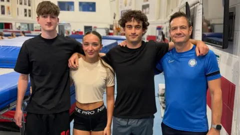 Four people standing in a gym, by trampolines, that are behind them. A man, wearing a black T-shirt and short is to the right, with short dark hair, then a woman, wearing black shorts, long hair tied back and a cream top. Mateo Jeannesson is wearing a black T-shirt, and blue shorts and has short curly hair, and Harvey Smith, a blue top, dark shorts, with short dark hair. 