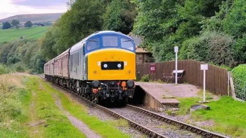 EastLancsRailway A yellow and blue train arrives at an empty platform in a rural area, with trees running along one side of the train and a grass bank on the other. Hills can be seen in the distance. 