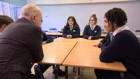 Teenage students grouped around desks listening to an adult