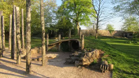 Bishops Palace A large play area built into a bank of grass. A dragon is coming out of the grass and there is a tunnel behind it. There are large wooden poles dotted around the play area.