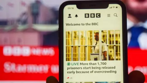 In this photo illustration, a BBC News website seen displayed on phone and TV screens