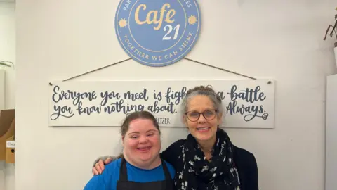 Two women stood in front of a sign for the cafe, one is wearing glasses. The sign reads: Cafe 21 Together we can shine.