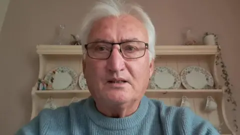 Peter Horgan has short white hair and glasses. He is wearing light blue sweater. Behind him is a cream‑coloured shelving unit filled with decorative plates