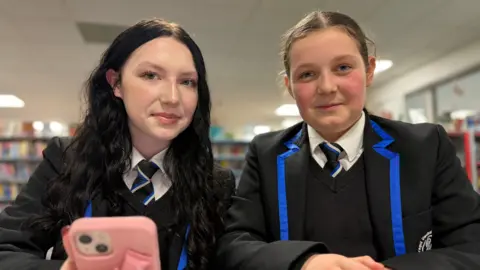 BBC Two girls wearing school uniform with black blazers and blue edging standing together in a library with one holding a pink mobile