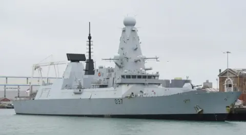 PA Media HMS Duncan, a grey battleship, in dock. It is a destroyer. Buildings can be seen on the dockside.