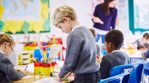 Getty Images A blonde haired boy in a grey uniform stands at a desk doing a craft. Some other students are sitting at a table. A teacher at the top of the class wearing a purple top looks at some of their work. 
