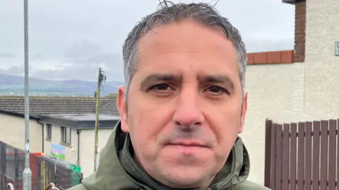 Christopher Jackson has short hair and is wearing a large green coat. He is standing near a house.