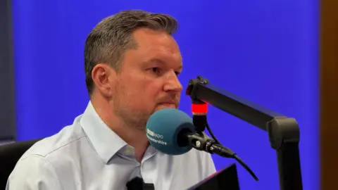 Kevin McNamara - a middle aged man with greying hair - sits in the BBC Radio Gloucestershire studio. He is wearing a pale blue shirt and is sat behind a microphone. There is a bright purple lightbox behind him.