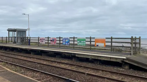 Across a railway line five information boards are attached to a wooden and wire fence. The boards are a variety of bright colours: pink, red, blue, green and orange. Further up the platform there is a glass shelter for travellers to wait in.