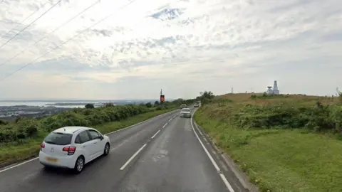 Cars driving on James Callaghan Drive in Portsmouth. It is a cloudy day. The sea can be seen in the distance.