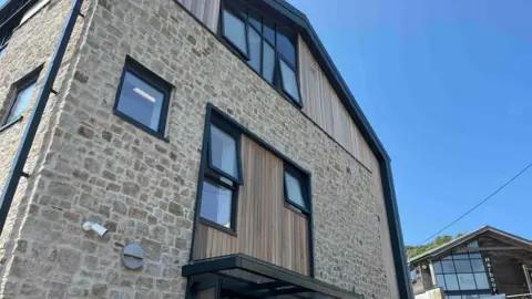 The image shows the exterior of a newly built three-storey stone and timber-clad building with large black-framed windows and a glass entrance canopy. Taken from ground level looking upwards, the photograph highlights the modern design of the structure against a clear blue sky, with another harbour-side building visible in the background.