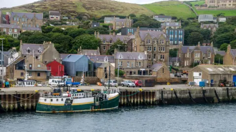 Getty Images Stromness harbour with a green and yellow boat tied up and buildings and cars in the background