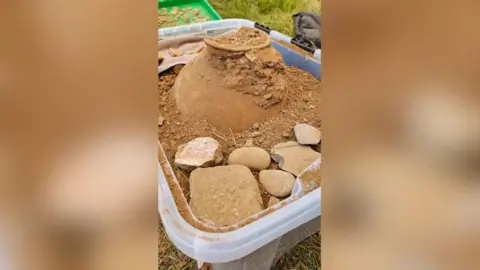 Minelab The top of a clay pot sticking out of an Ikea box surrounded by dirt and stones.