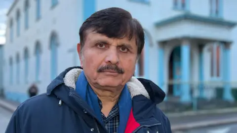 Sarfaraz is wearing a dark winter coat stands on a quiet street in front of a large pale-blue and white building with arched windows. 