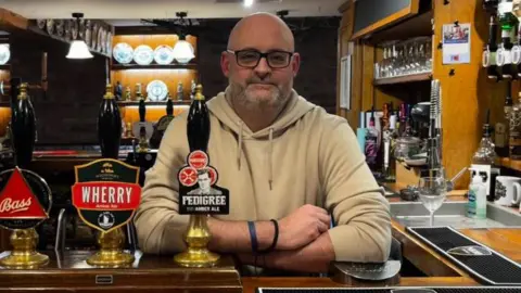 Lewis stood behind the bar of the pub. He is wearing a beige hoodie, black glasses and has his arms folded.